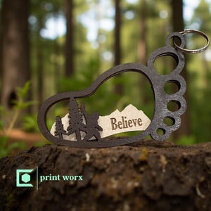 May include: A dark brown wooden keychain in the shape of a footprint, featuring a cutout design with a Bigfoot silhouette, trees, and the word "Believe." The keychain has a silver keyring attached.