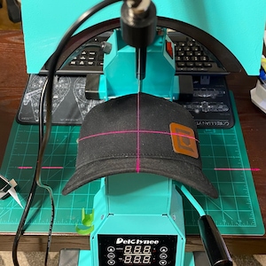 May include: A teal heat press machine with a digital display labeled DelClynee, featuring a laser alignment guide projecting a pink crosshair onto a black baseball cap. The cap has a brown rectangular leather patch on the front panel.