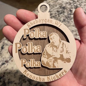 May include: Wooden Christmas ornament with a circular design. The ornament features the words "Sheboygan, WI" and "Kenosha Kickers" in a circular pattern. The word "Polka" is repeated three times. A portrait of a man is in the center.