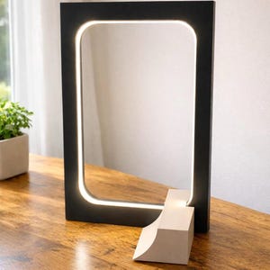 May include: A modern black rectangular LED lamp with a white light border. The lamp has rounded corners and sits on a wooden surface. A small, off-white geometric base supports the lamp.