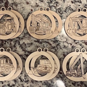Könnte beinhalten: Zehn Holzornamente mit verschiedenen Baufahrzeugdesigns. Jedes kreisförmige Ornament hat ein ausgeschnittenes Design eines Bulldozers, Baggers und anderer Maschinen. Die Ornamente sind aus hellem Holz gefertigt und haben oben ein kleines Loch zum Aufhängen.