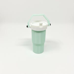 Puede incluir: Un vaso de color verde menta con tapa blanca y asa verde claro. El vaso tiene forma cil&iacute;ndrica con una base ligeramente m&aacute;s ancha. El asa est&aacute; sujeta a los lados de la tapa con herrajes negros.