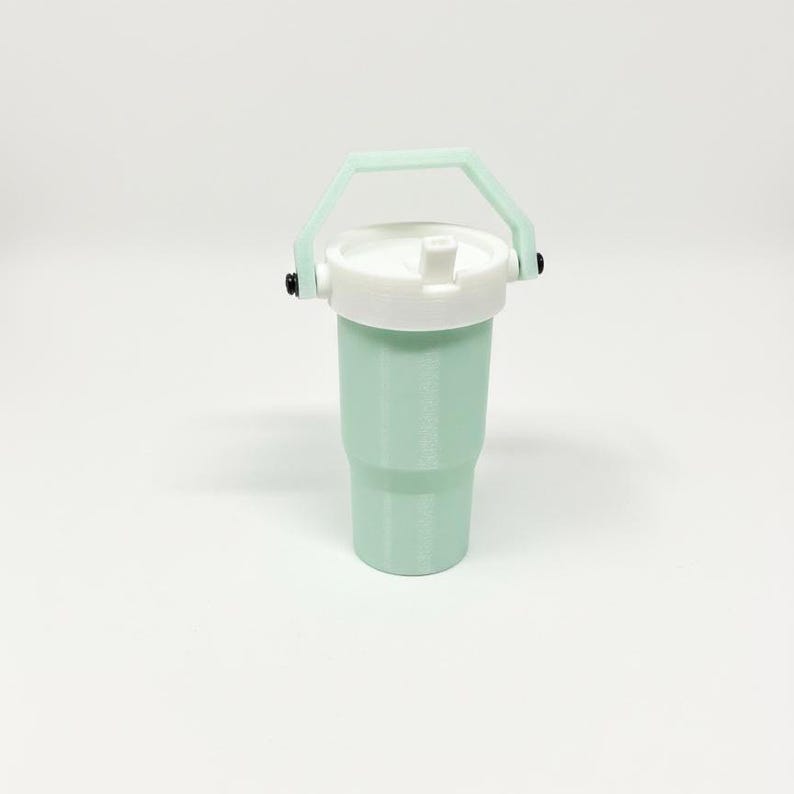 Puede incluir: Un vaso de color verde menta con tapa blanca y asa verde claro. El vaso tiene forma cil&iacute;ndrica con una base ligeramente m&aacute;s ancha. El asa est&aacute; sujeta a los lados de la tapa con herrajes negros.