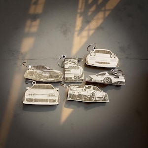 May include: A collection of car-shaped keychains, each with a silver keyring. The keychains are made of a light-colored material and feature detailed car designs. The cars include various models, with visible headlights and body details.