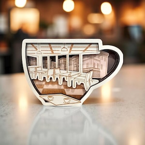 Könnte beinhalten: Ein dekoratives Objekt aus Holz in Form einer Kaffeetasse mit einem mehrschichtigen Design. Das Innere zeigt eine Cafészene mit Tischen, Stühlen und Gebäudefassaden. Die Tasse ist hellbraun mit weißem Umriss.