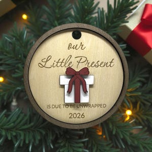 Pode incluir: Enfeite de Natal redondo em madeira com borda castanha escura e centro castanho claro. O texto "our Little Present" está gravado acima de uma caixa de presente branca com um laço vermelho. O texto "IS DUE TO BE UNWRAPPED 2026" está abaixo.