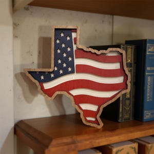 May include: A wooden Texas-shaped wall art piece featuring the American flag design. The art piece has a natural wood frame, a blue section with white stars, and red and white stripes. It is displayed on a wooden shelf.