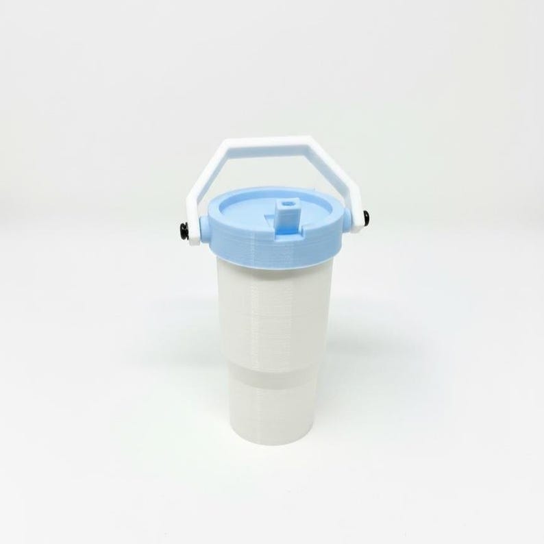 Puede incluir: Un vaso blanco con tapa y asa azul claro. La tapa tiene una abertura abatible. El asa es blanca y est&aacute; sujeta al vaso con bisagras negras. El vaso est&aacute; dise&ntilde;ado para contener bebidas.