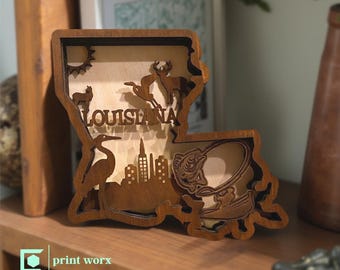 May include: A wooden Louisiana-shaped decorative piece with laser-cut details. The design includes a sun, animals, the word "LOUISIANA", a cityscape, and a fishing scene. The piece is brown and sits on a wooden shelf.