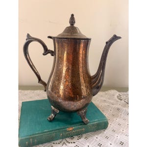 May include: A tarnished silver-plated coffee pot featuring a curved handle, a long spout, and a lid topped with a finial. The body of the pot has a bulbous base supported by three ornate feet, showing significant oxidation and dark patina throughout.