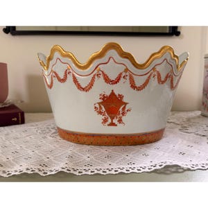 Puede incluir: Un cuenco de cerámica blanca con un borde festoneado en dorado. El exterior presenta guirnaldas florales pintadas en naranja y un motivo central de urna naranja. La base está decorada con una banda naranja con un patrón de puntos.