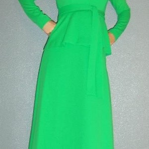 Puede incluir: Un vestido largo de color verde con cuello alto y cintur&oacute;n. El vestido tiene un corpi&ntilde;o ajustado y una falda fluida. La modelo lleva el vestido con un par de tacones negros.