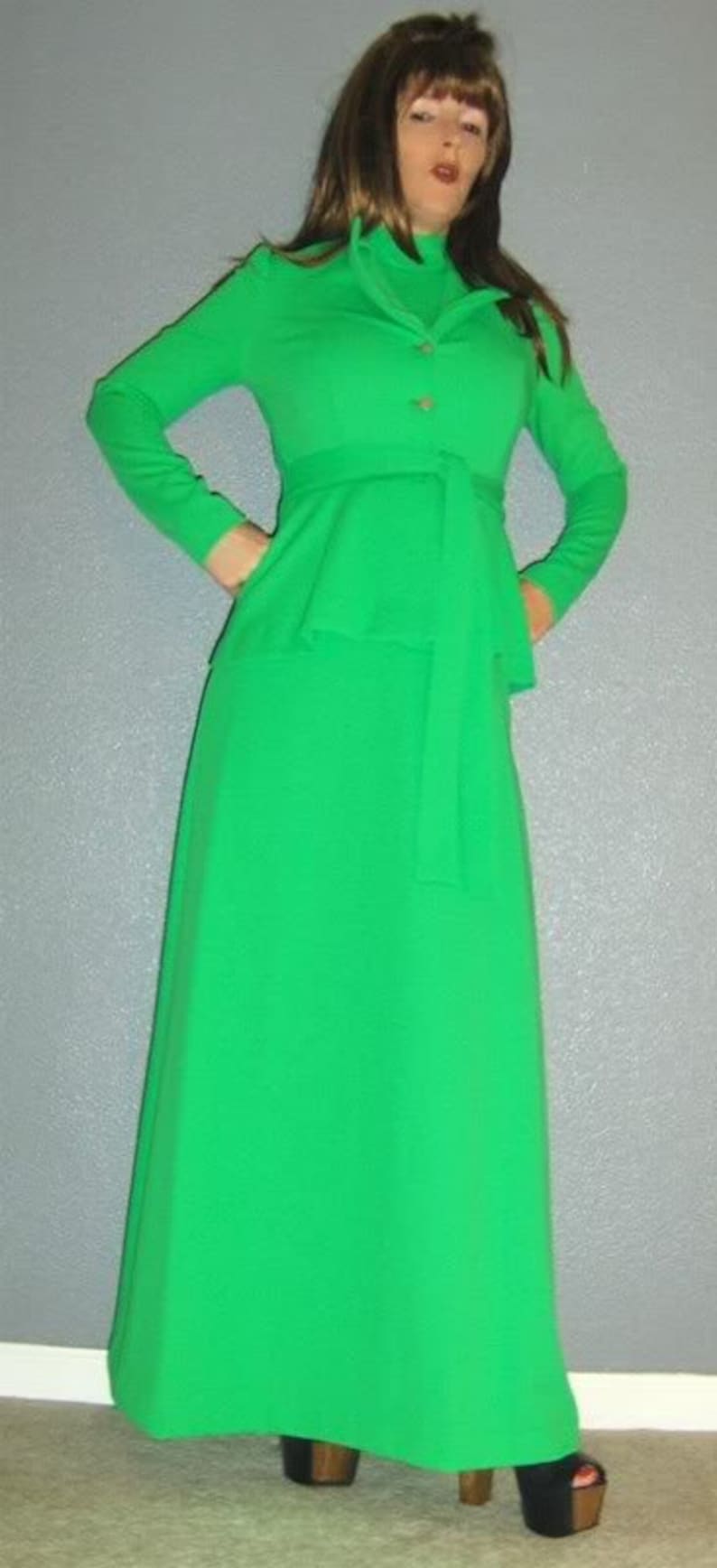 Puede incluir: Un vestido largo de color verde con cuello alto y cintur&oacute;n. El vestido tiene un corpi&ntilde;o ajustado y una falda fluida. La modelo lleva el vestido con un par de tacones negros.