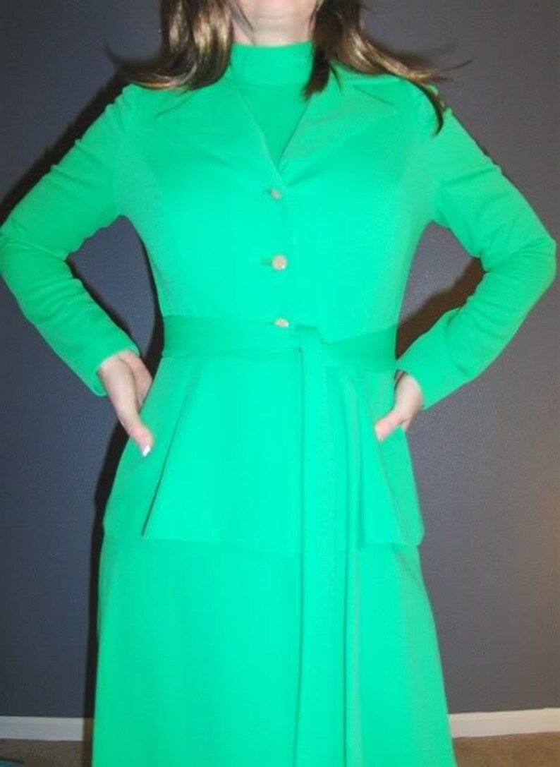 Puede incluir: Un vestido largo de manga larga, de color verde, con cuello alto, cierre de botones y cintur&oacute;n. El vestido tiene un corpi&ntilde;o ajustado y una falda fluida.