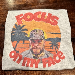 May include: A gray t-shirt featuring a graphic with the text "FOCUS ON MY FACE" in red. The graphic includes a man wearing a hat, palm trees, and a sunset design in orange and yellow.