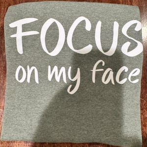 May include: A heather green t-shirt with the phrase "FOCUS on my face" printed in white, bold, cursive lettering. The shirt is folded and lies on a wooden surface.