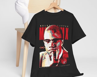 May include: Black t-shirt featuring a portrait of Malcolm X with the text "Malcolm X" in red and white. The image includes the years 1925-1965. A white baseball cap rests on a wooden chair in the background.