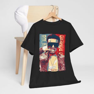 May include: Black t-shirt featuring a graphic of a man wearing sunglasses, with the text "ROLA ROMERO" in red and blue vertical stripes. The shirt is hanging on a wooden hanger.