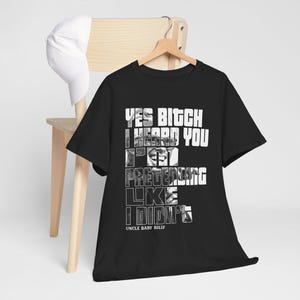 以下が含まれることがあります： 「YES BITCH I HEARD YOU I'M PRETENDING LIKE I DIDN'T」の白い文字がプリントされた黒いTシャツ。木製のハンガーにかけられています。白い野球帽が木製の椅子の上に置かれています。