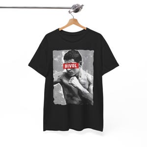 May include: Black t-shirt featuring a grayscale image of a boxer with the name "BIVOL" in red block letters across his eyes. The design has a distressed, textured background.