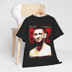 May include: Black t-shirt featuring a stylized portrait of Khamzat Chimaev with the number 72 in red. The text "Khamzat Chimaev" and "1994" are printed at the bottom. A white baseball cap hangs on a wooden chair.