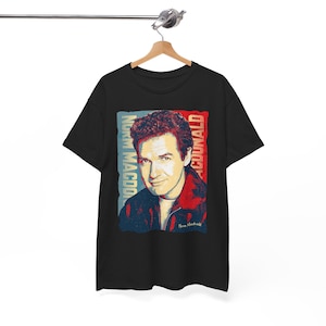May include: Black t-shirt featuring a portrait of Norm Macdonald with the text "Norm Macdonald" in blue and "Macdonald" in red. The portrait is in a pop art style, with a vintage aesthetic.