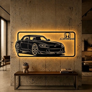 Puede incluir: Arte mural de metal negro que representa una silueta de coche Honda S2000 con el logotipo S2000. La obra de arte está iluminada con un brillo amarillo cálido, montada en una pared de hormigón texturizado. Debajo hay una mesa de madera con una pequeña planta y libros.