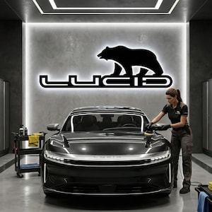 May include: A black Lucid electric vehicle being detailed in a modern garage. The Lucid logo, featuring a bear silhouette, is illuminated on the wall. A person is polishing the car's hood. The garage has cabinets and tools.