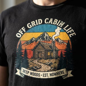 Op de afbeelding: Een zwart t-shirt met een grafische print van een blokhut in het bos met bergen op de achtergrond. De tekst luidt OFF GRID CABIN LIFE bovenaan en DEEP WOODS EST. NOWHERE op een banner onderaan, in een kleurenpalet van een zonsondergang.