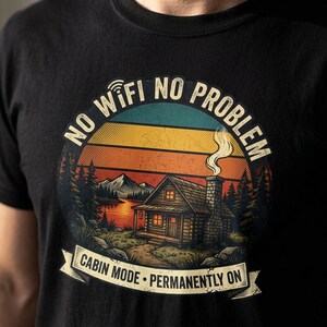 May include: A black t-shirt featuring a circular graphic of a log cabin by a lake with mountains in the background. The text reads NO WIFI NO PROBLEM at the top and CABIN MODE PERMANENTLY ON at the bottom, set against a retro sunset sky.
