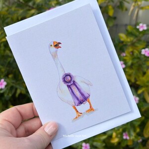 May include: A white greeting card featuring a watercolor illustration of a white goose with an orange beak and feet, wearing a purple award ribbon around its neck. The text Michelle Jaye Art is printed in small white letters at the bottom center.