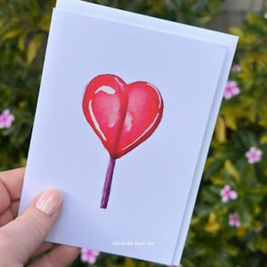 May include: A white greeting card featuring a watercolor illustration of a red heart-shaped lollipop with a purple stick. The text Michelle Jaye Art is printed in small white letters at the bottom center of the card surface.