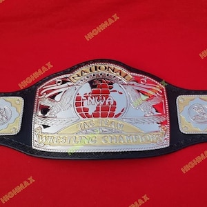 May include: A wrestling championship belt with a black leather strap and a silver and gold metal center plate. The plate features the text "NATIONAL NWA TAG TEAM WRESTLING CHAMPIONS" and a globe design.