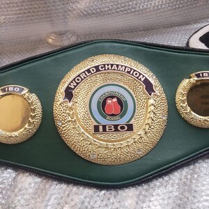 Pu&ograve; includere: Una cintura da campione di boxe in pelle verde con medaglioni dorati. Il medaglione centrale recita "World Champion" e presenta una grafica di guantoni da boxe. La cintura &egrave; etichettata "IBO".