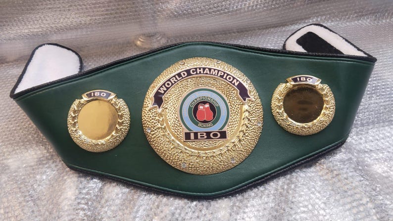 Pu&ograve; includere: Una cintura da campione di boxe in pelle verde con medaglioni dorati. Il medaglione centrale recita "World Champion" e presenta una grafica di guantoni da boxe. La cintura &egrave; etichettata "IBO".