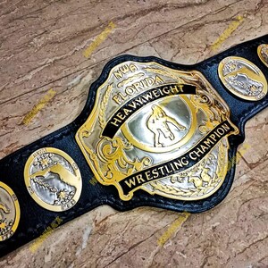 May include: A black leather wrestling championship belt with gold and silver detailing. The belt has a large central plate with the words "NWA FLORIDA HEAVYWEIGHT WRESTLING CHAMPION" and smaller plates with state imagery.