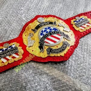 Könnte beinhalten: Ein roter und goldener Wrestling-Meisterschaftsgürtel mit den Worten "United States Champion" und "IWGP". Der Gürtel hat ein zentrales Emblem mit dem Design der amerikanischen Flagge und goldfarbenen Akzenten.