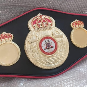 Pu&ograve; includere: Una cintura da campionato di boxe nera con dettagli dorati e rossi. La cintura presenta tre medaglioni dorati con dettagli a corona rossa. Il medaglione centrale presenta un emblema circolare con testo e figure di pugilato.