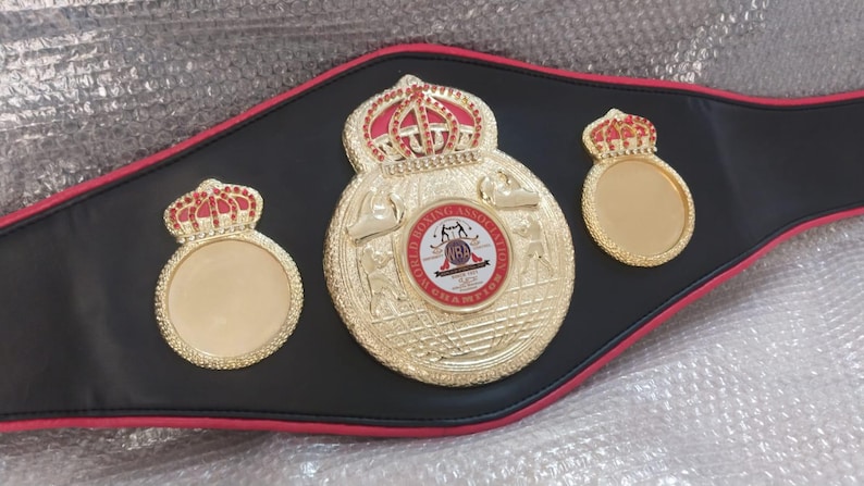 Pu&ograve; includere: Una cintura da campionato di boxe nera con dettagli dorati e rossi. La cintura presenta tre medaglioni dorati con dettagli a corona rossa. Il medaglione centrale presenta un emblema circolare con testo e figure di pugilato.