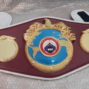 Pu&ograve; includere: Una cintura da campione di boxe in pelle bordeaux con dettagli dorati. Il centro presenta un globo blu con il logo WBO e un'aquila dorata. Due medaglioni dorati affiancano il centro. Imbottitura bianca e nera.
