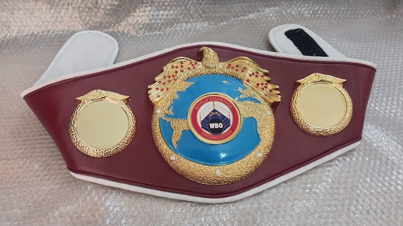 Pu&ograve; includere: Una cintura da campione di boxe in pelle bordeaux con dettagli dorati. Il centro presenta un globo blu con il logo WBO e un'aquila dorata. Due medaglioni dorati affiancano il centro. Imbottitura bianca e nera.