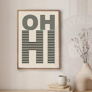 May include: A framed art print with the words "OH HI" in a bold, minimalist design. The letters are black and composed of horizontal lines, set against a cream background. The frame is a light brown color. A vase with dried flowers and a coffee cup are on a wooden surface.