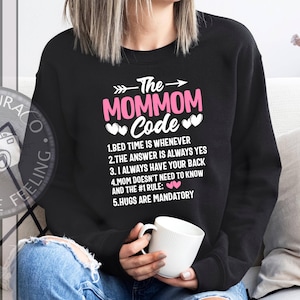 May include: Black sweatshirt with the text "The MOMMOM Code" in pink and white. The design includes an arrow, hearts, and a list of rules. The wearer is holding a white mug.