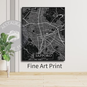May include: A monochrome fine art print of Sapporo, Japan, city map. The map shows detailed street layouts and is labelled "Sapporo - Japan". The print is displayed on a wall with a plant in a white pot.