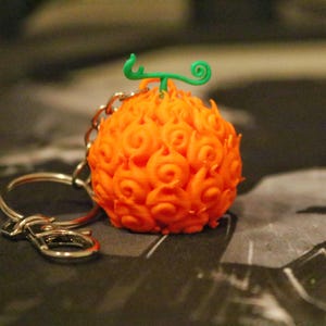 Puede incluir: Un llavero naranja brillante con forma de fruta, con un tallo verde y un llavero plateado. El colgante tiene una superficie texturizada con un patrón de llamas. El llavero es probablemente de plástico.