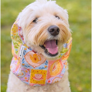 May include: A light-colored dog wearing a fabric neck gaiter featuring a colorful patchwork pattern of teapots, teacups, and floral motifs in shades of yellow, pink, and blue. A small white tag with the text Coppy Paws is visible on the side.
