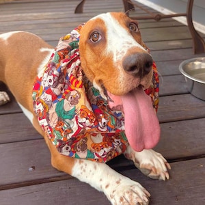 May include: A Basset Hound with a long pink tongue, wearing a colourful bandana featuring cartoon dogs. The dog is lying on a wooden deck near a metal bowl.