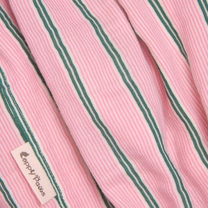 Puede incluir: Una tela con patr&oacute;n de rayas finas rosas y blancas que presenta rayas verticales de color verde oscuro. Una peque&ntilde;a etiqueta rectangular de tela color hueso est&aacute; cosida al material, mostrando el texto "Coppy Paws" en una fuente estilizada marr&oacute;n oscuro.