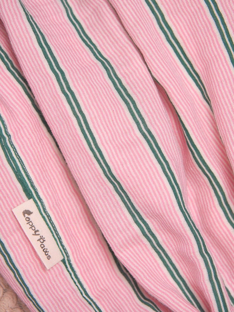 Puede incluir: Una tela con patr&oacute;n de rayas finas rosas y blancas que presenta rayas verticales de color verde oscuro. Una peque&ntilde;a etiqueta rectangular de tela color hueso est&aacute; cosida al material, mostrando el texto "Coppy Paws" en una fuente estilizada marr&oacute;n oscuro.