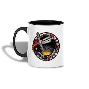 Puede incluir: Taza de cerámica blanca con asa e interior negros. La taza presenta un diseño circular con un cohete, el texto "PROJECT HAIL MARY" y varias banderas internacionales. El diseño está sobre un fondo negro con un sol naciente.